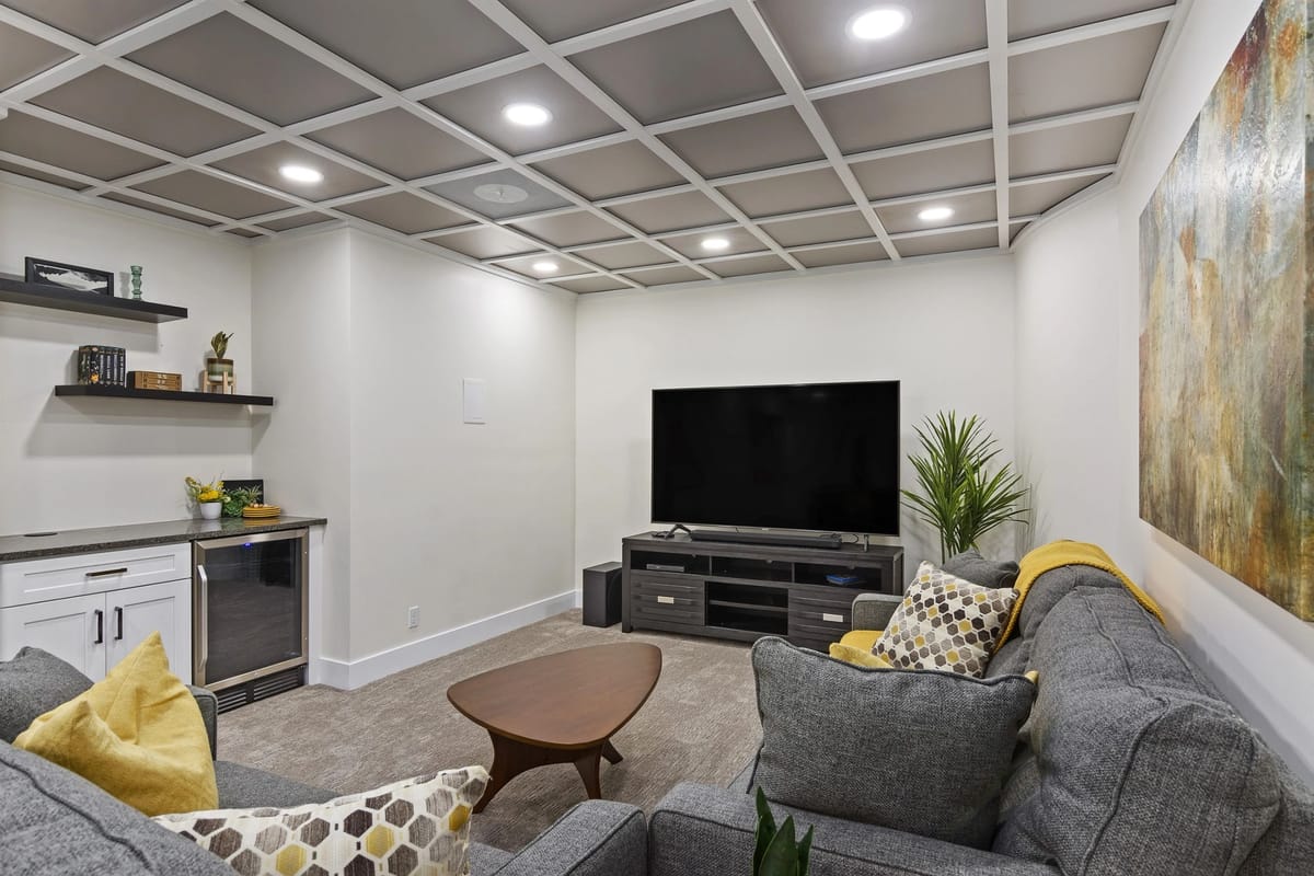 Finished basement family room with a grey coffered ceiling, recessed lighting, a wall-mounted television, a sectional sofa and a built-in wet bar with beverage fridge and floating shelves.