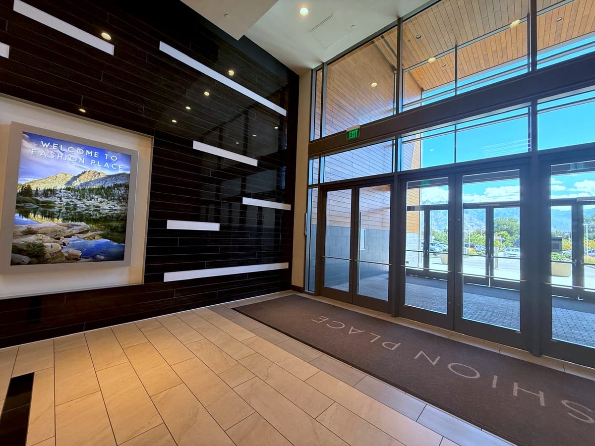 Entry vestibule inside Fashion Place: a dark linear-tile wall with an illuminated Welcome to Fashion Place lightbox on the left, full-height glazed entrance doors under a cedar-clad ceiling on the right, and a recessed walk-off mat set flush into the tile floor.