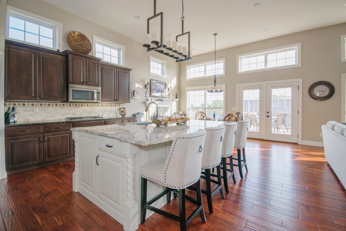 Open-concept great room with a large white kitchen island seating four, dark stained perimeter cabinetry, hardwood floors, two-storey windows and French doors leading onto a deck.